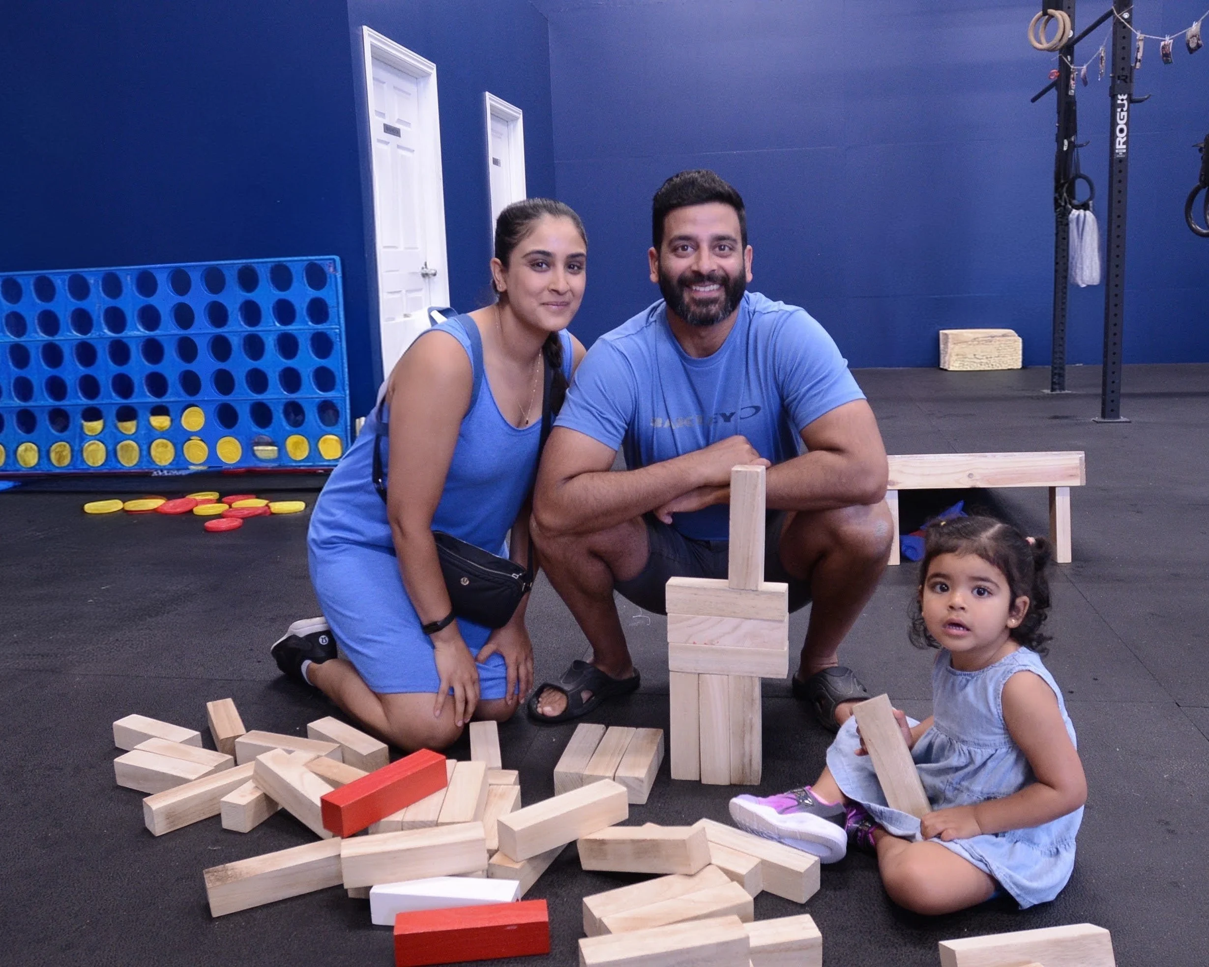 Two adults and a child, all smiling and dressed in blue, are in a room with gym equipment and giant boardgames 
