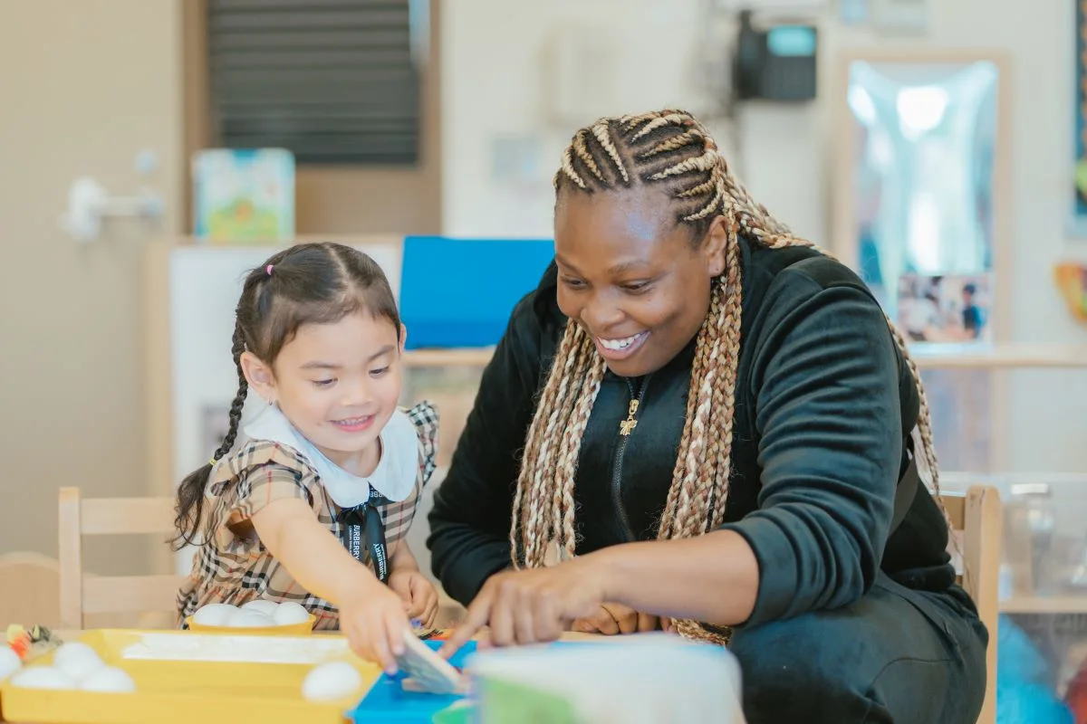 A smiling Y M C A educator, sitting beside a young child at a table in a child care room, points to a colourful activity they are exploring.