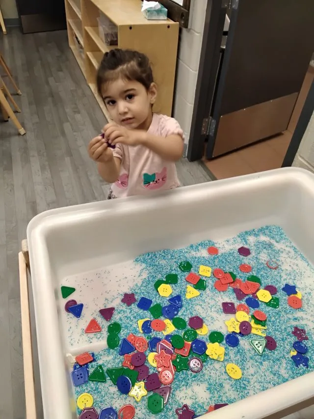 A toddler wearing a pink shirt is playing with a colourfu shapes in a binl 