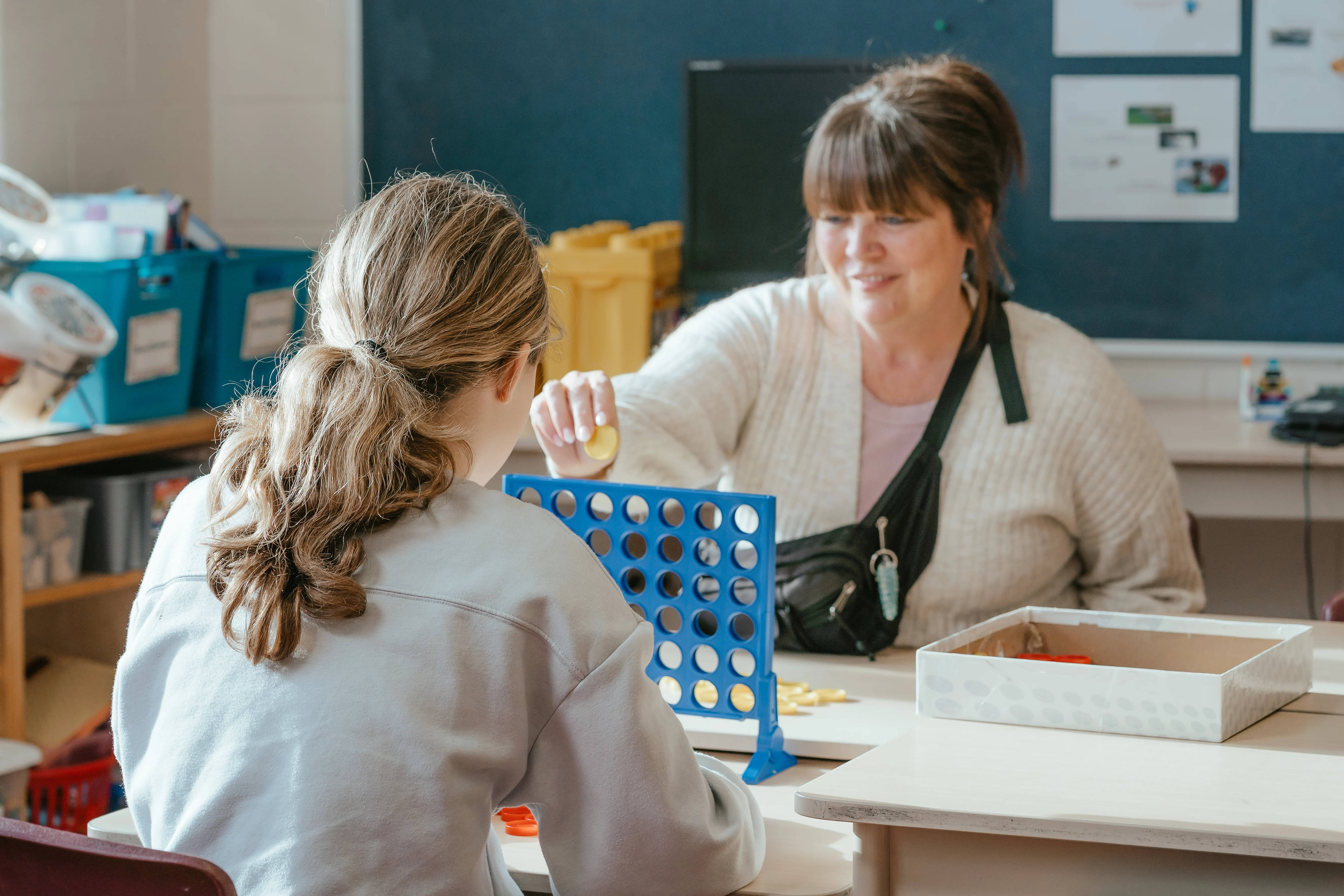 A child care educator in a white sweater and a child in a grey sweatshirt play a boardgame in a child care centre classroom