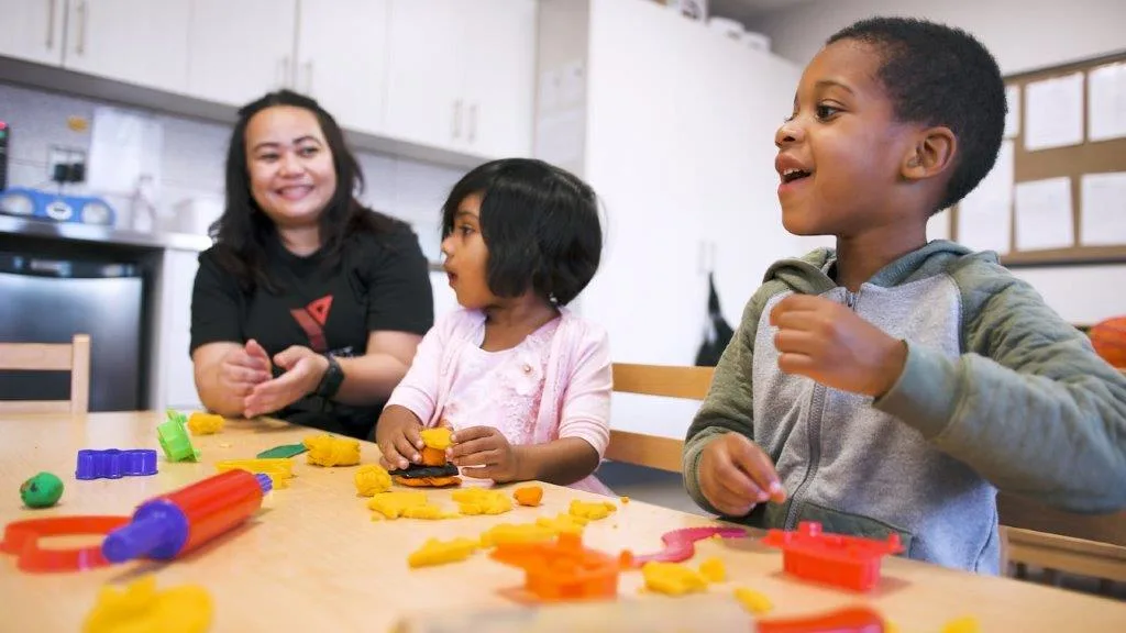 A child care educator and two young children sit at table cutting out shapes from plasticene with cookie cutters