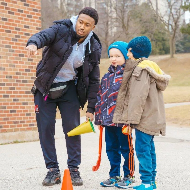 A child care educator holding pylons instructs two children in a school yard. 