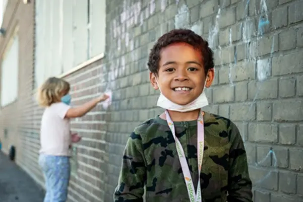a boy with a mask on, smiling, while a child in the background draws with chalk on the wall