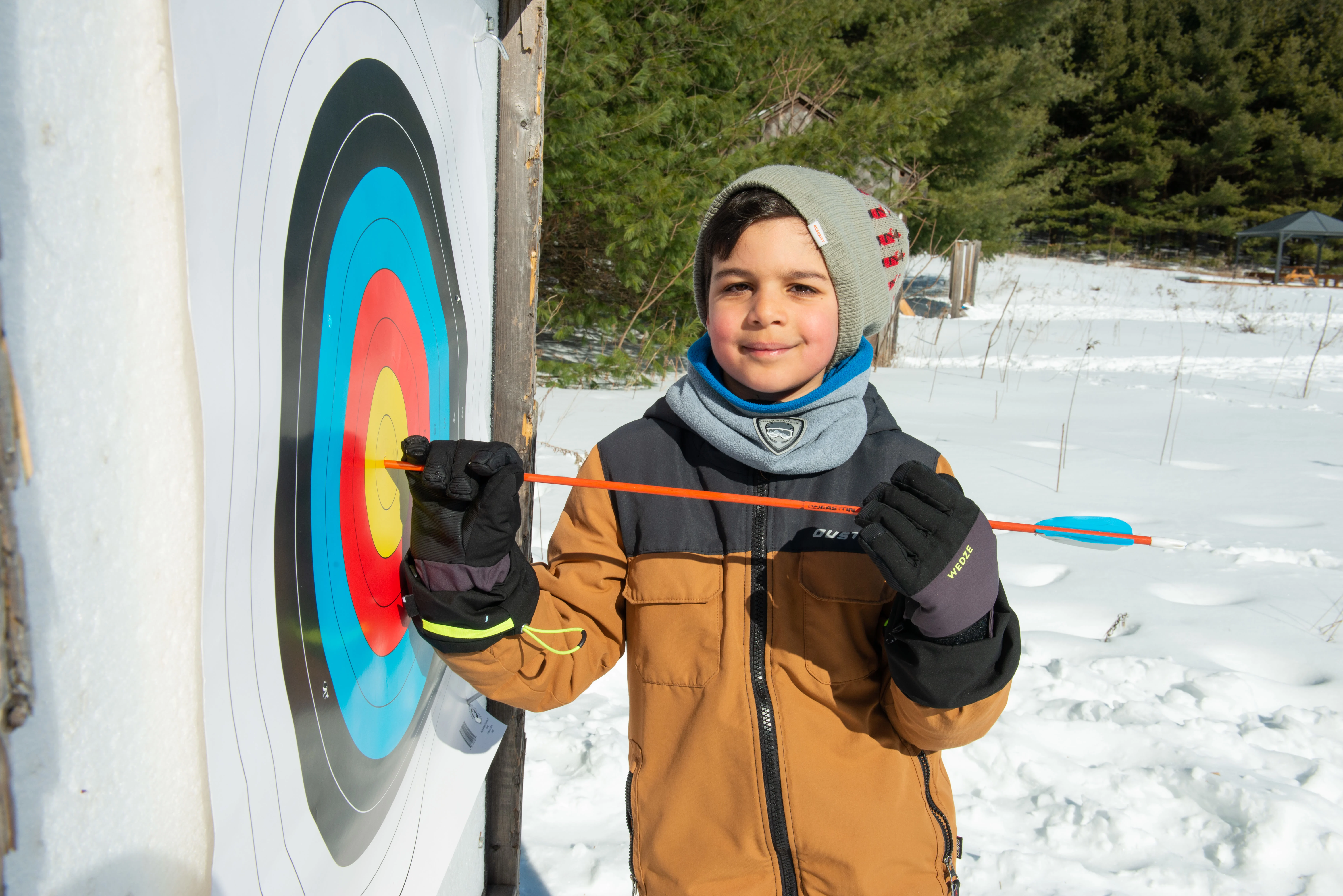 Young camper smiling at camera wearing winter clothing pulling an arrow out of a archery target.  Snow is in the background.