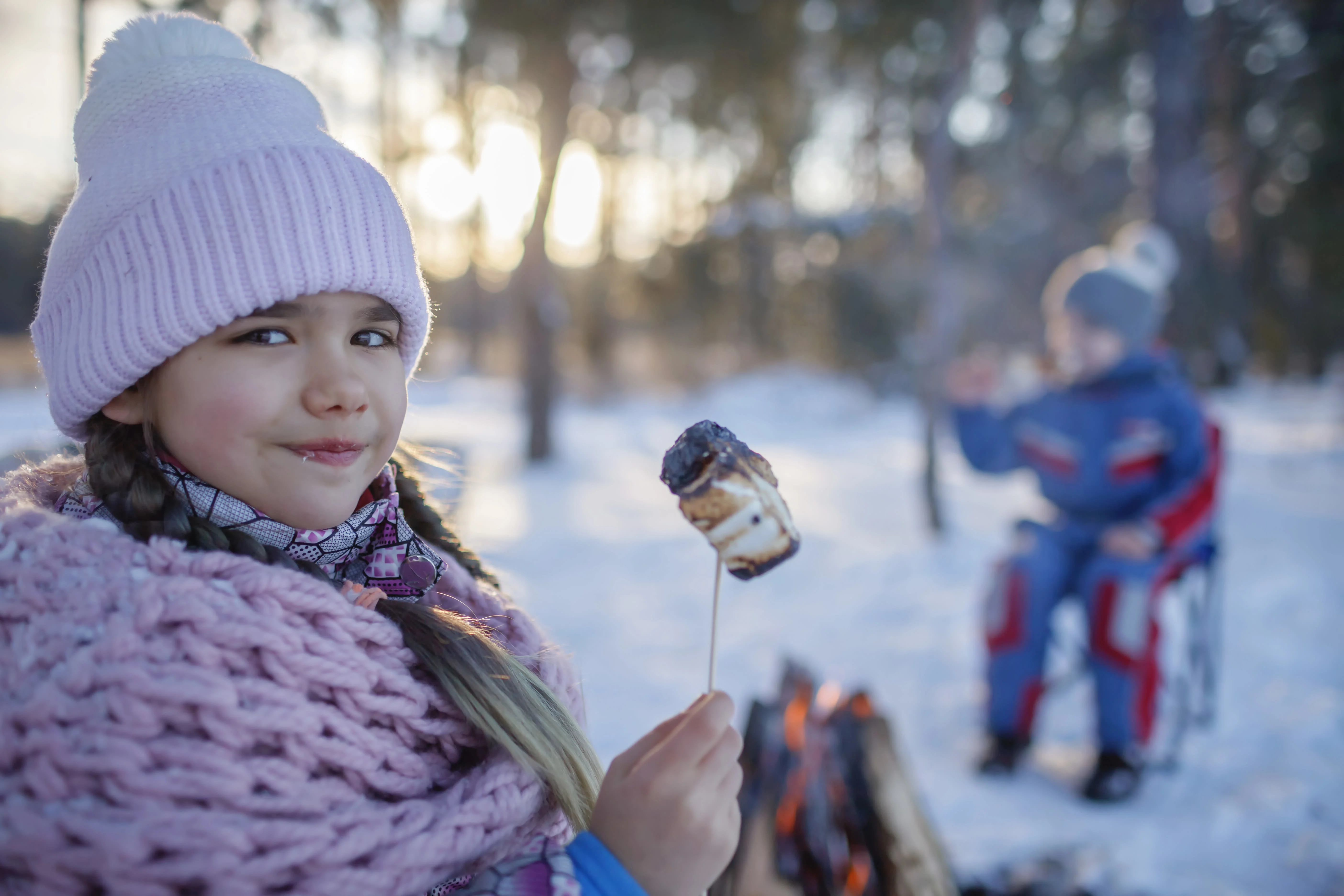 Young camper outside in winter clothing roasting a marshmellow with a fire and snow behind them