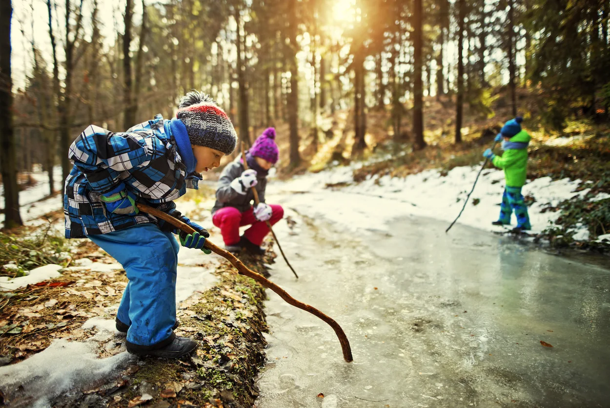 Three kids aged 10 and 6 are playing with a huge frozen puddle in winter forest. They are breaking ice on the puddle with sticks
