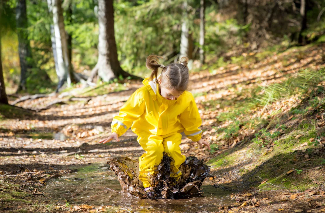 Cute little girl jumping in muddy puddle wearing yellow rubber overalls. Happy childhood. Sunny autumn forest