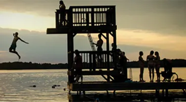 a silhouette of a group of kids jumping off a diving board in the lake