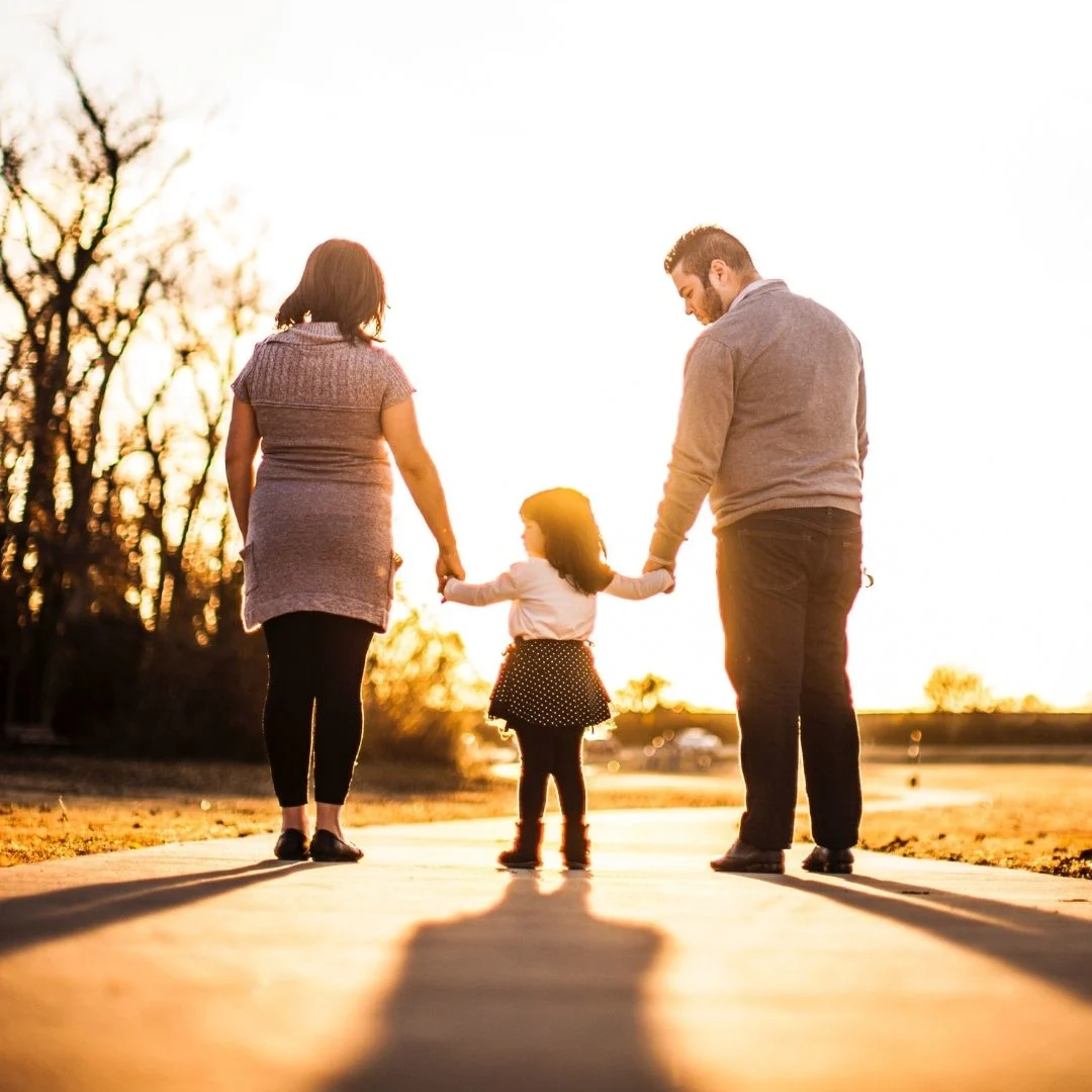backlit family of 3 with child in the middle, holding parents' hands