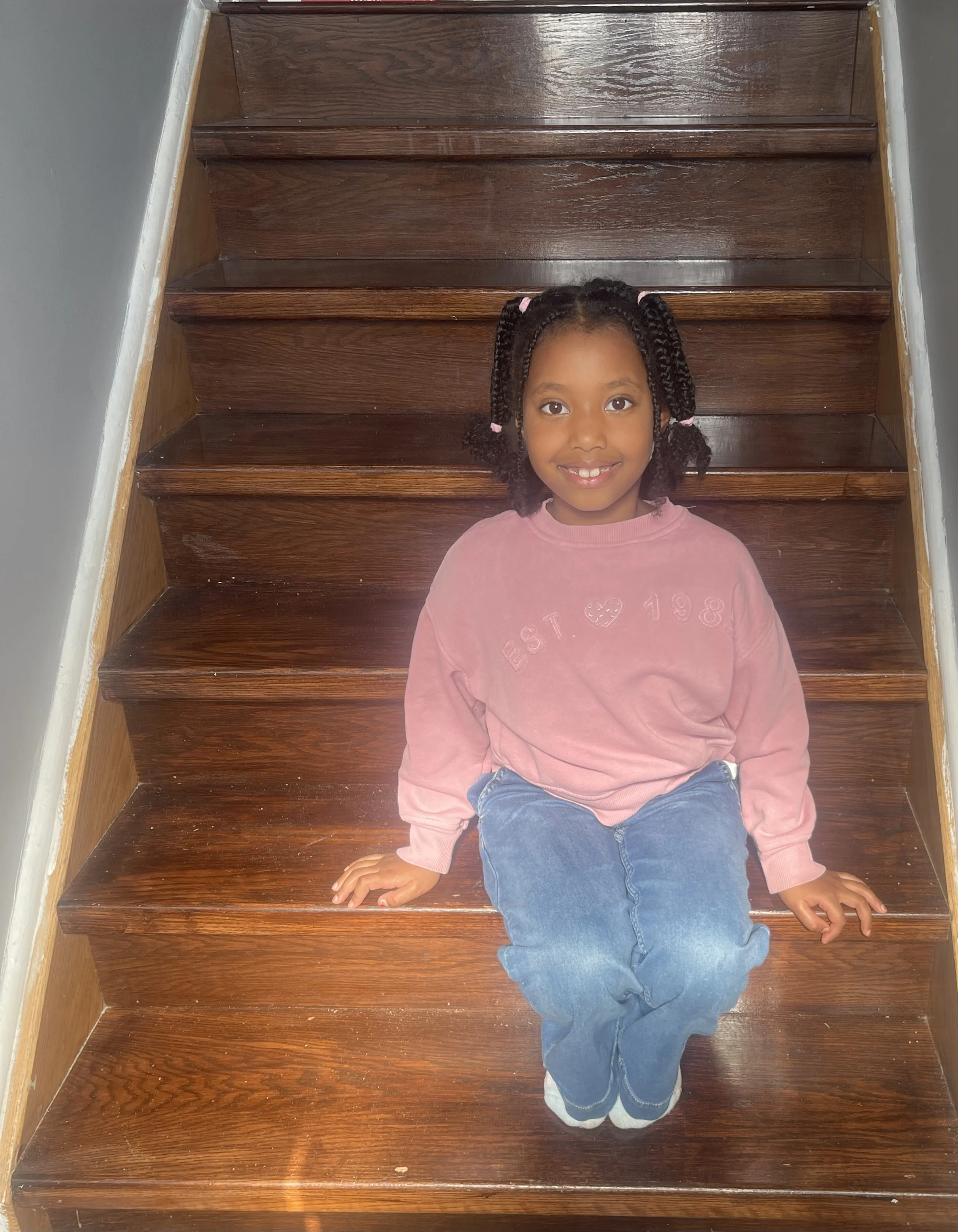 A child with braided hair in a pink sweatshirt sits on a staircase smiling.