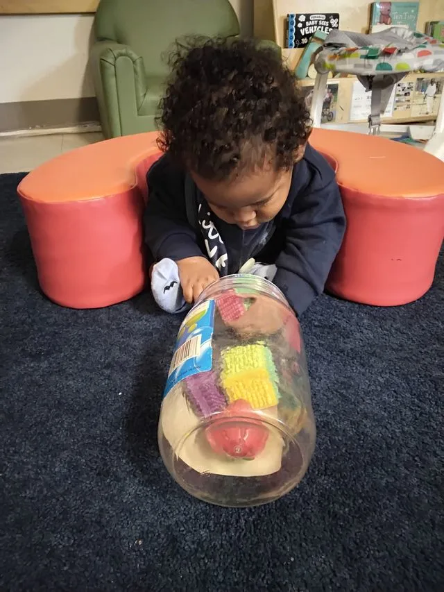 Small child sitting on carpet, leaning forward playing with colourful items in a clear jar.