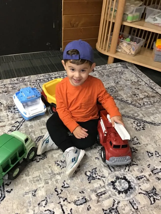 A child in a blue cap and orange shirt sits on a rug happily playing with toy trucks
