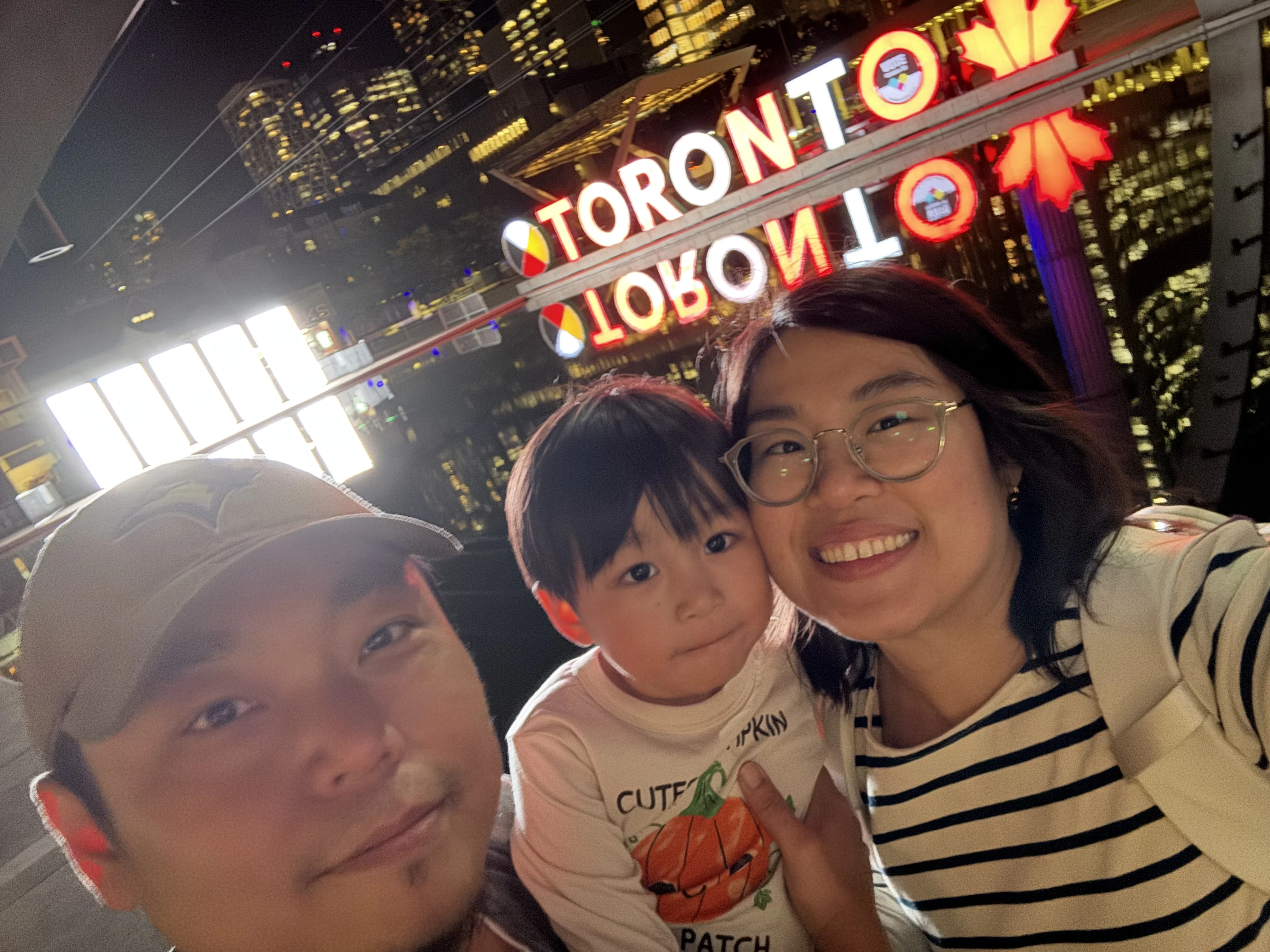 A young child sits between two adults. They are in front of neon sign that says Toronto.