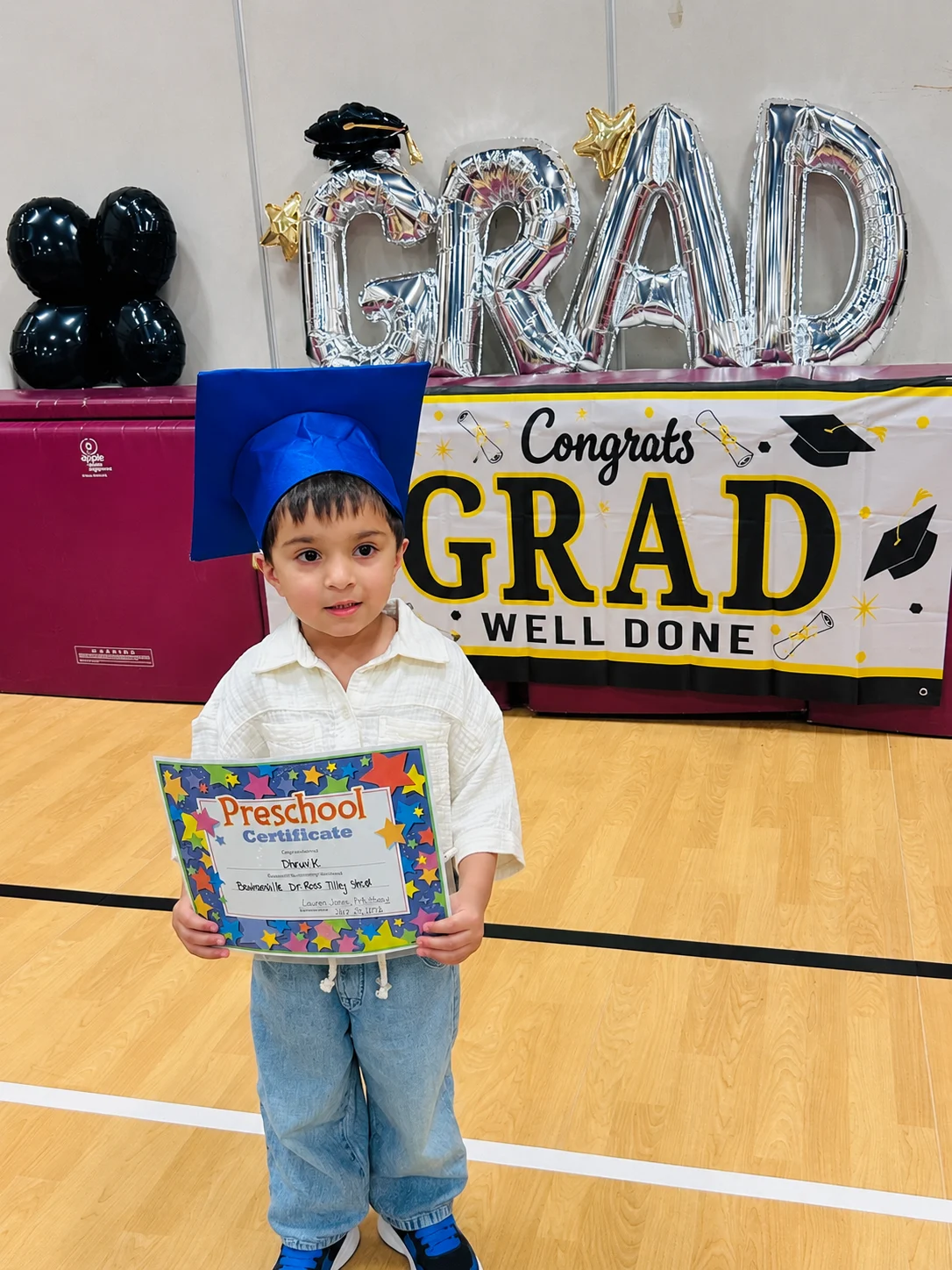 A young child in a blue graduation cap holds a certificate and stands in front of a banner that reads Congrats Grad Well Done