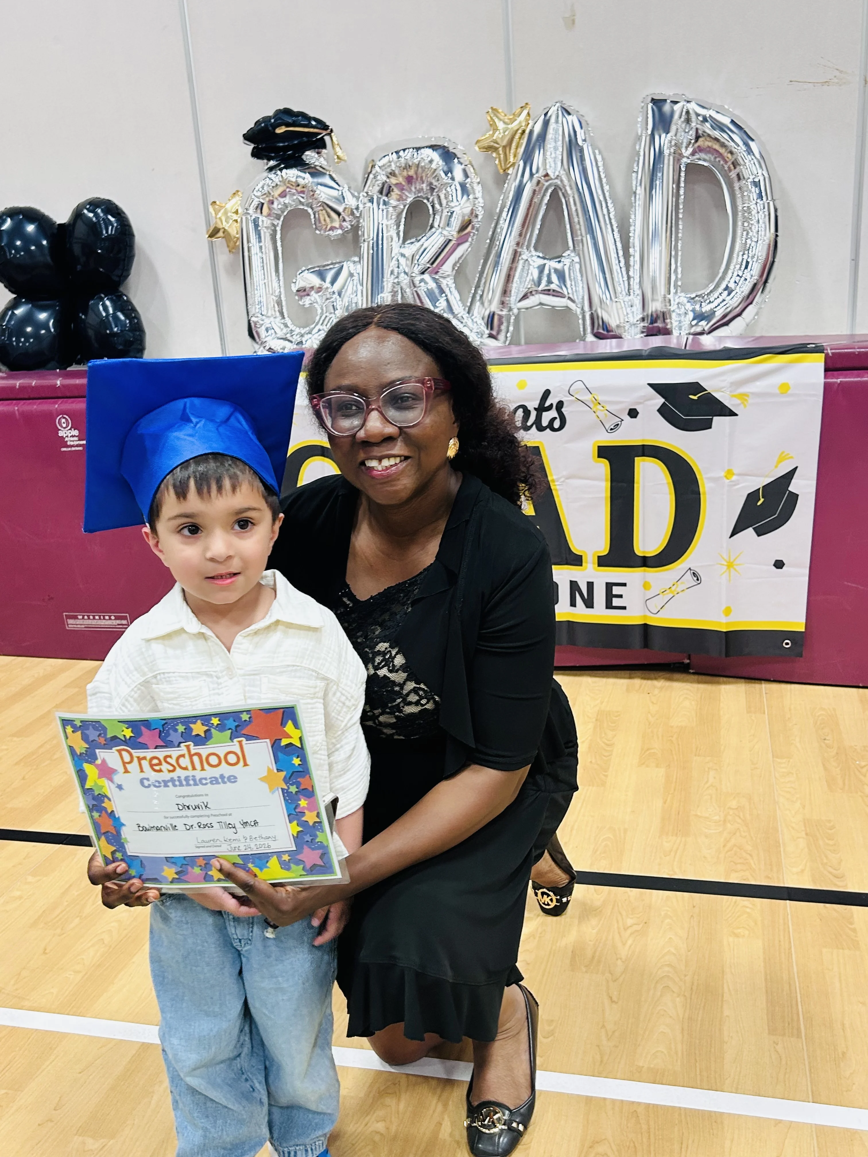 A young child in a blue graduation cap holds a certificate while sitting on the knee of a smiling adult with dark hair and glasses
