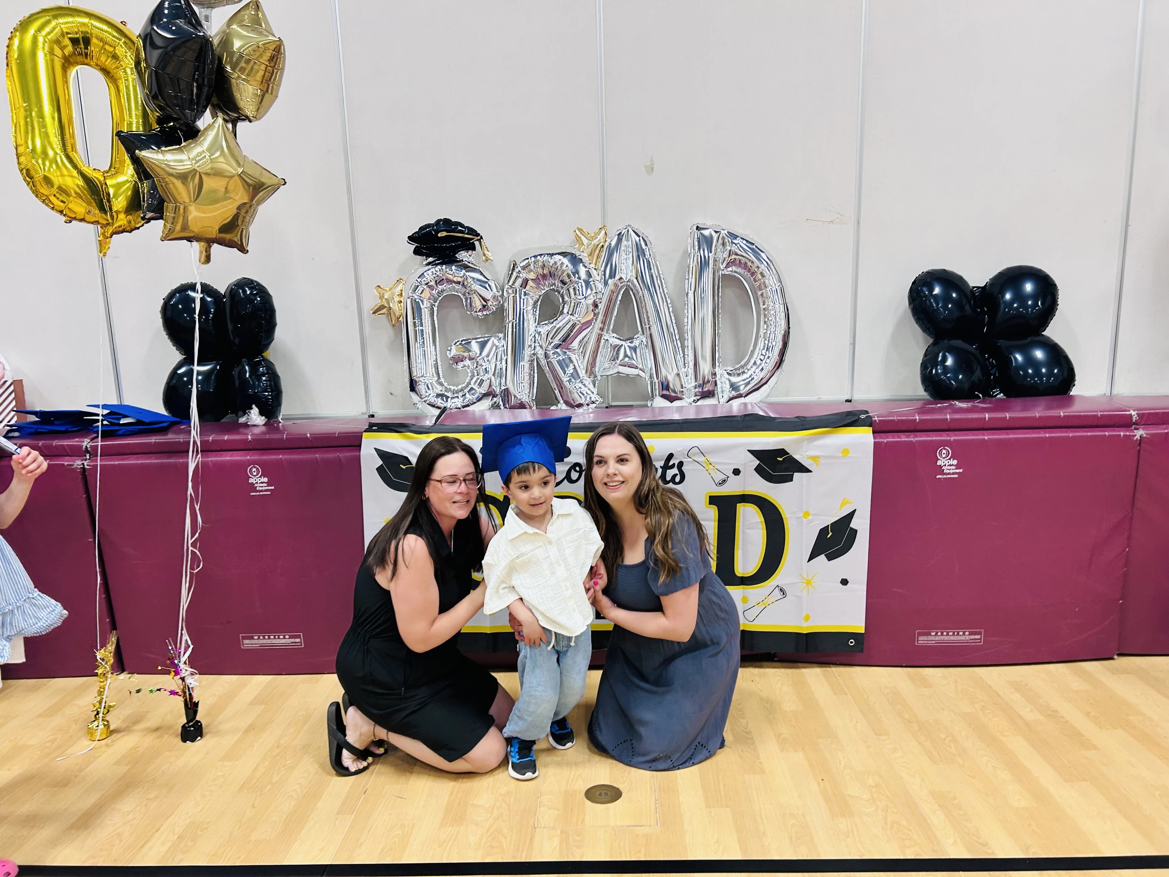 A young child in a blue graduation cap is flanked by two kneeling adults. Silver balloons spelling out the word GRAD are behind them