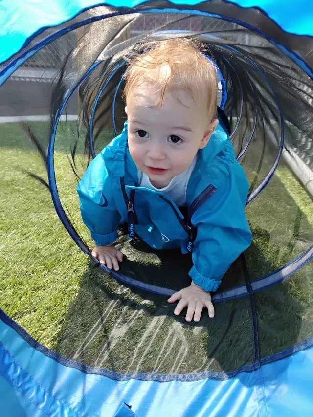 A smiling infant in a blue jacket crawls through an obstacle course tube in a playground.