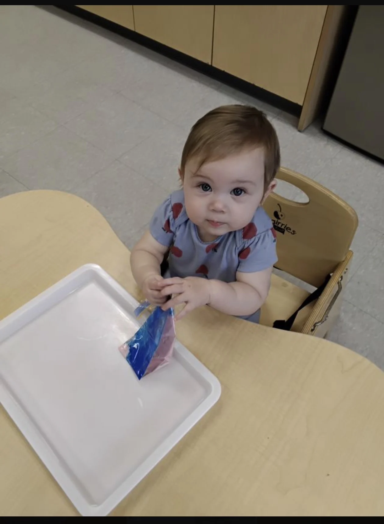 An infant sitting at a low table plays with a piece of blue paper on a white plastic tray.