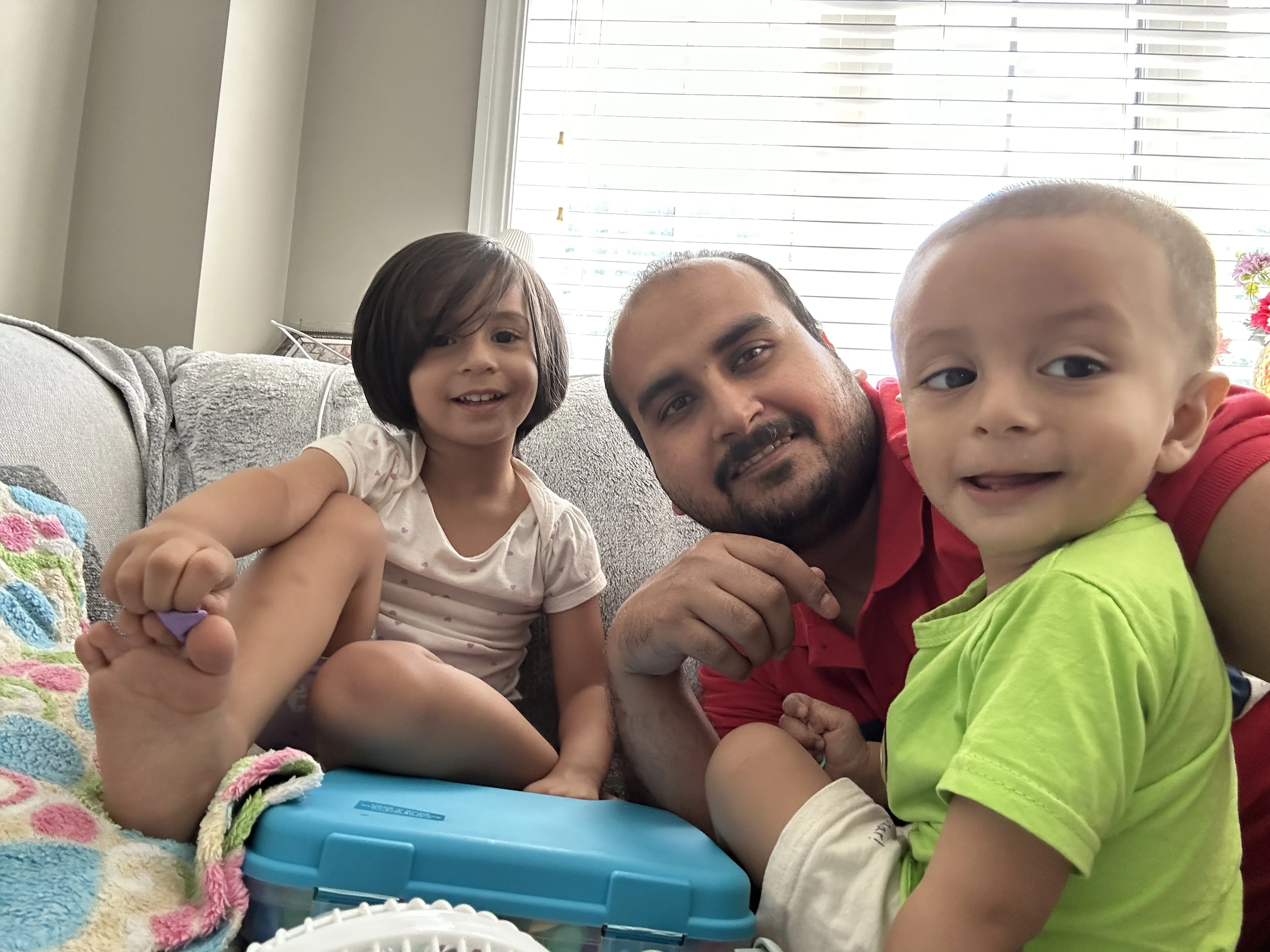 An adult in a red shirt and two young children sit closely together on a couch