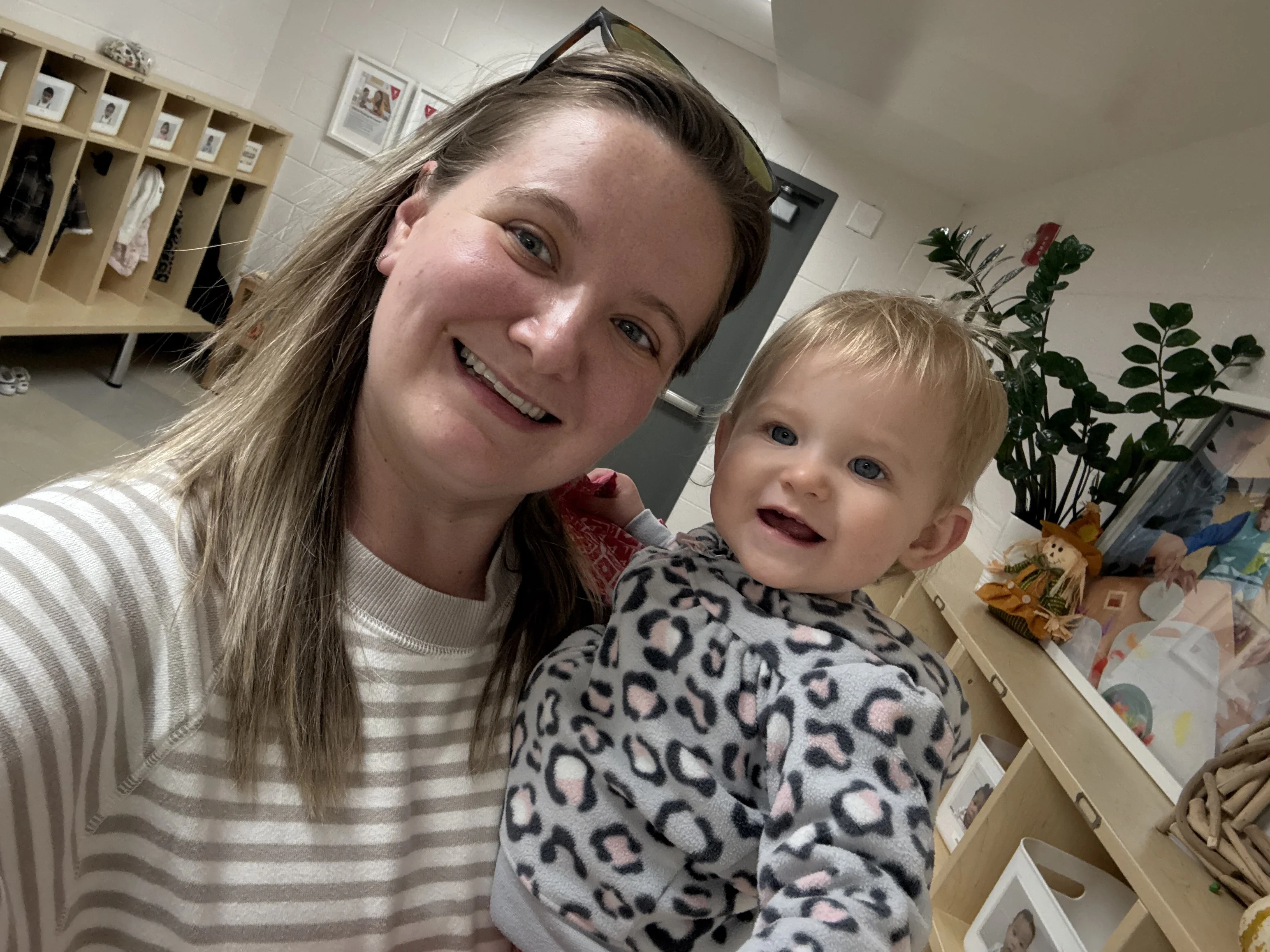 An adult with blond hair and sunglasses on their head is smiling and lifting a small child with blond hair and leopard print sweater. They stand in front of cubbies at a child care centre
