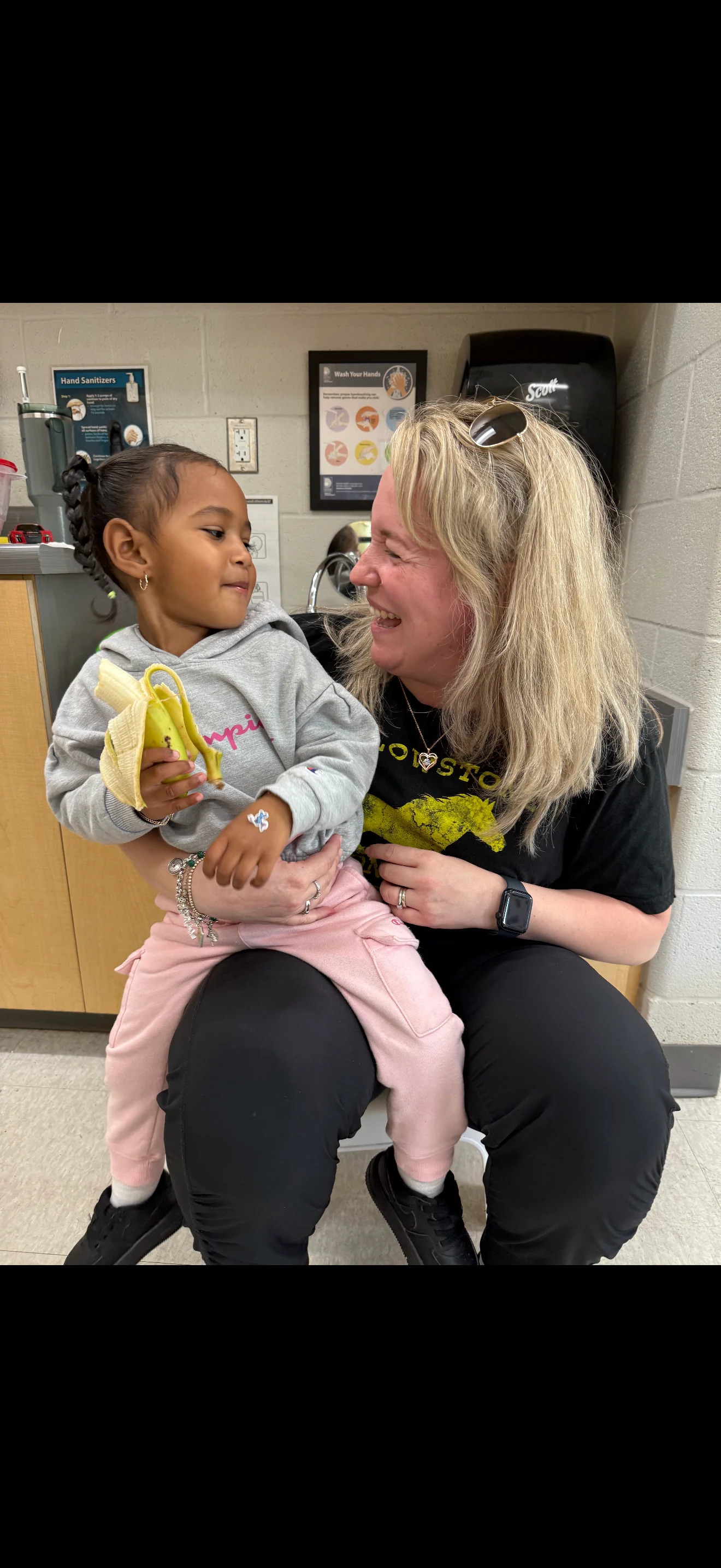 Young child sits on educator's knee eating a banana.
