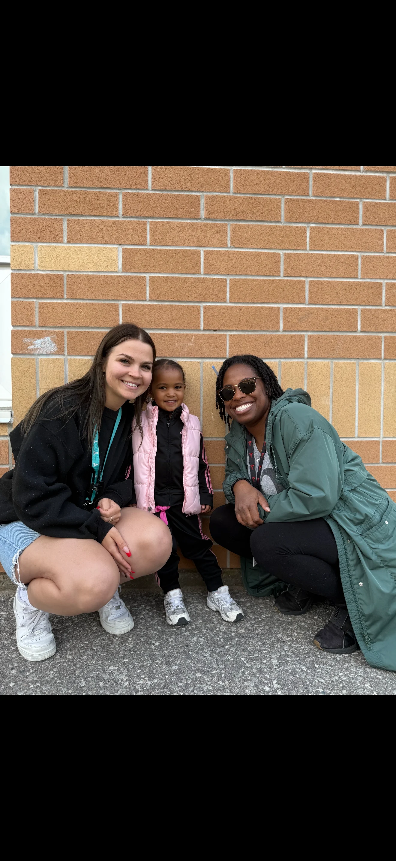 Two adults kneel against a brick all outside with a young child standing between them.