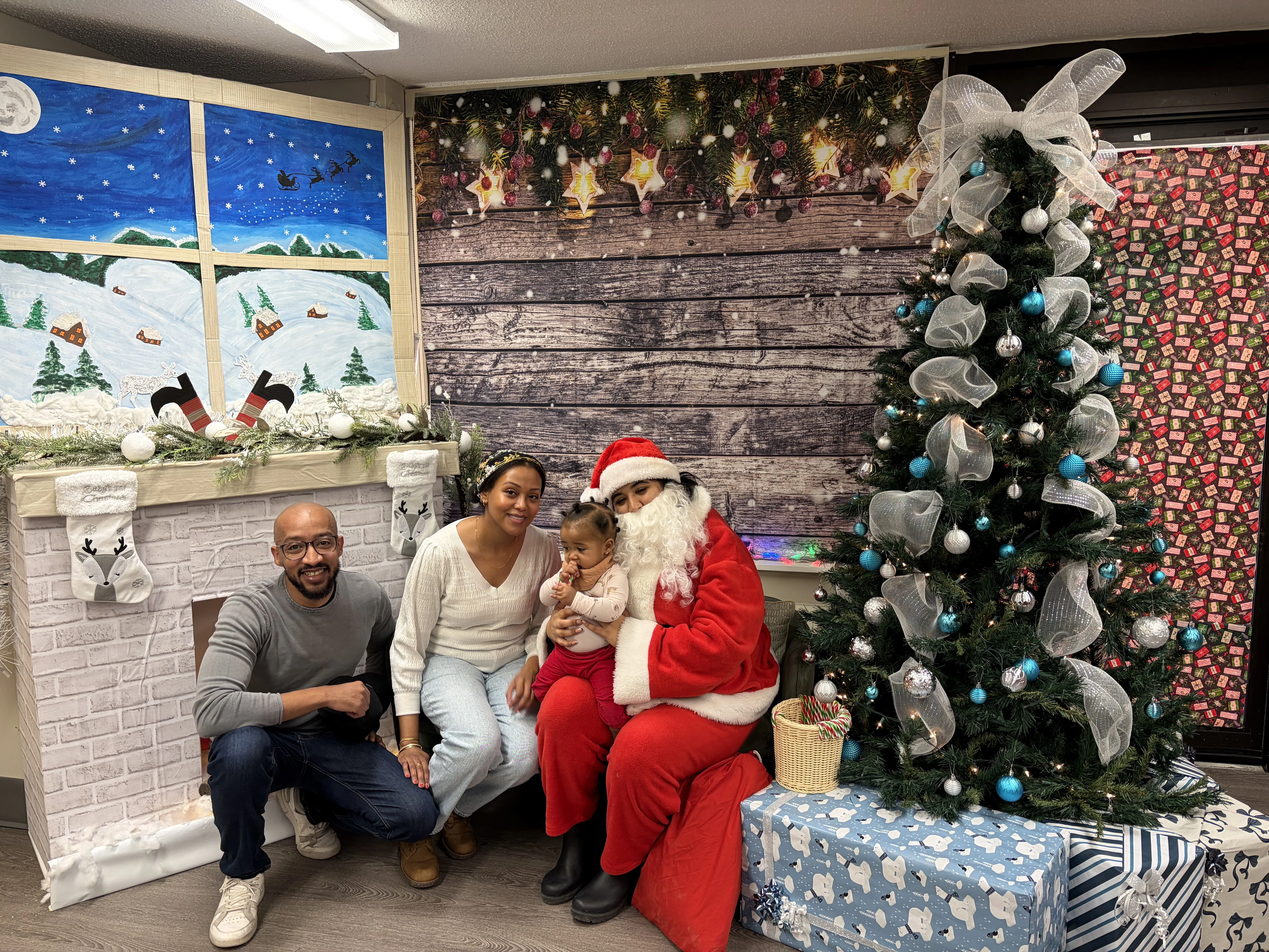 An infant in a white sweater sit on the lap of a person dressed as Santa Clause. Two smiling adults sit crouch beside them. They are in a festive 