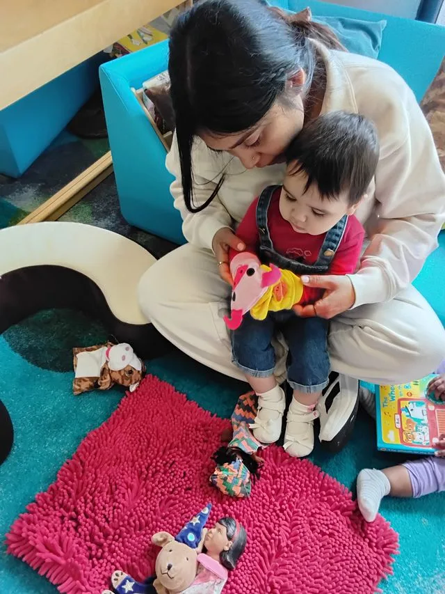 An educator and young child sit on the floor of a child care. The child has sock pockets on their hands.