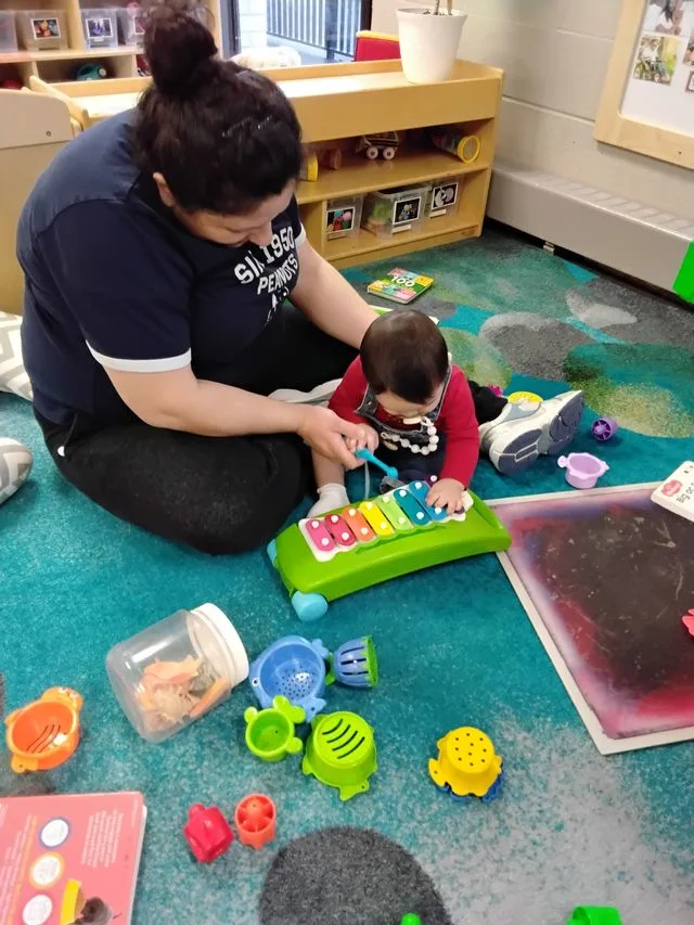 Educator and young child sit on the floor in a child care centre playing with an xylophone.