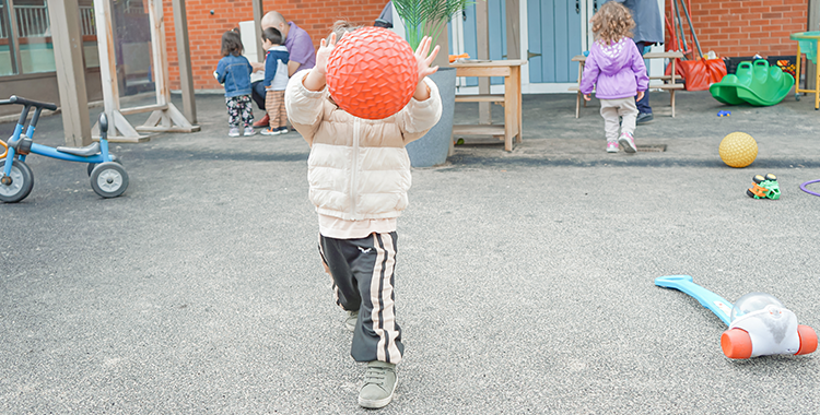 Children and educators play outdoors at the North York YMCA Child Care playground. A child throws a red ball.