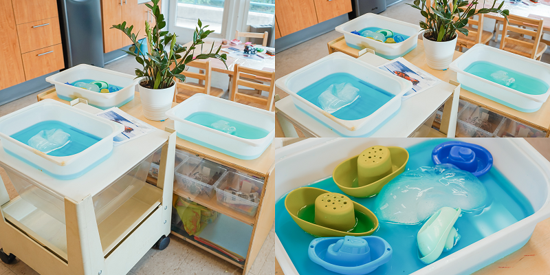 A toy boat programming setup at North York YMCA Child Care centre that includes three white plastic tubs with blue solution, an ice block, and 5 colourful toy boats in blue and green. 