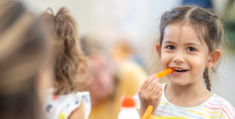 Children are seen sitting around a table eating their lunch. They are dressed casually and have healthy eating choices on the table in front of them as they talk amongst themselves.