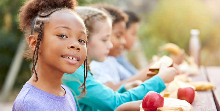 A child sits at an outdoor picnic table with three other children. They eat a healthy lunch including apples, bananas, and sandwiches.