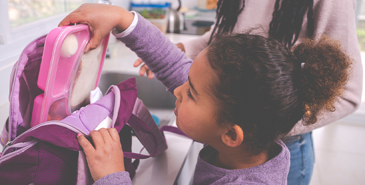 6 years old child packing a lunch box into a backpack. Their parent is in the background.