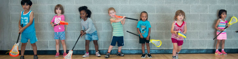 Children lined up against the gym wall with lacrosse sticks ready to play lacrosse.