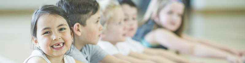 A young girl smiles at the camera while her classmates stretch in the background